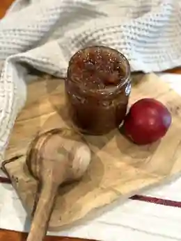 Borcan cu gem de nectarine pe tocător din lemn, alături de o nectarină și o lingură de lemn